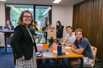 Smiling professor stands beside students working on laptops during a classroom activity at Bethel.