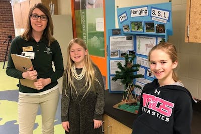 Kristi Berg ’07 stands with two students at a school in front of a posterboard presentation. 