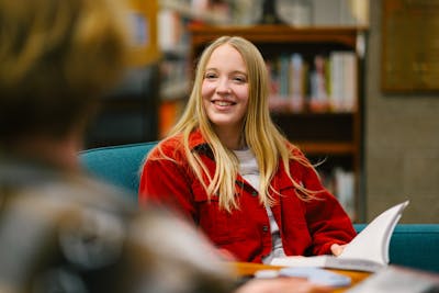 A student in a red jacket is smiling while discussing in a library. 