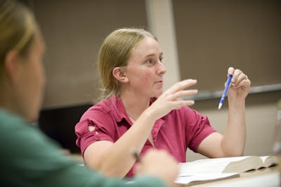 A student in a red shirt making a point during a classroom discussion, holding a pen.