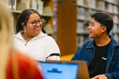 Bethel University students working together in a library, sharing a light moment while working on laptops.