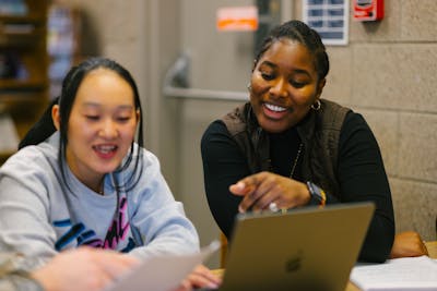 Two Bethel University students collaborating on a project, smiling and pointing at a laptop screen. 