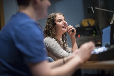 Bethel University students in a media production room, one wearing a headset and speaking into a microphone.