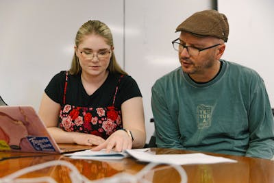 A Bethel University student and professor discussing a document, sitting at a table with a laptop and papers. 