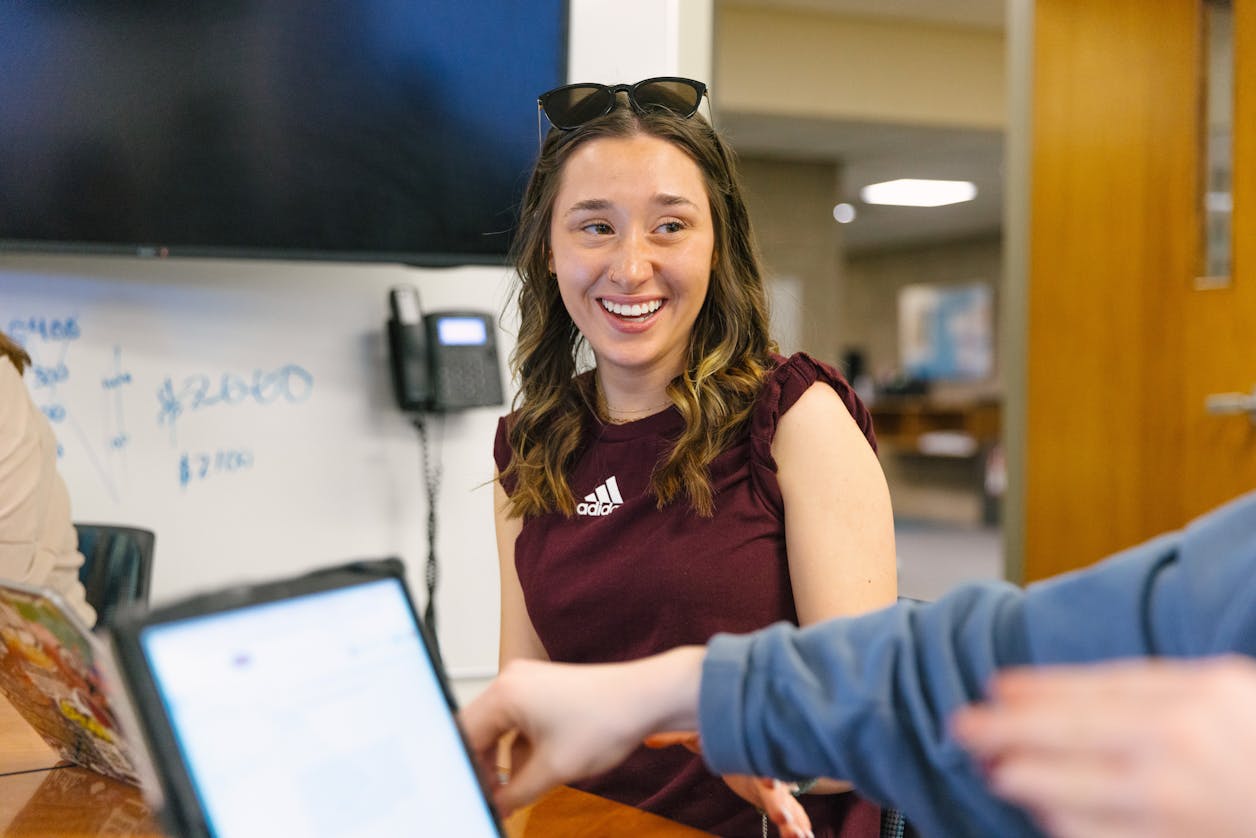 A student wearing a maroon Adidas shirt is smiling brightly during a classroom discussion. 