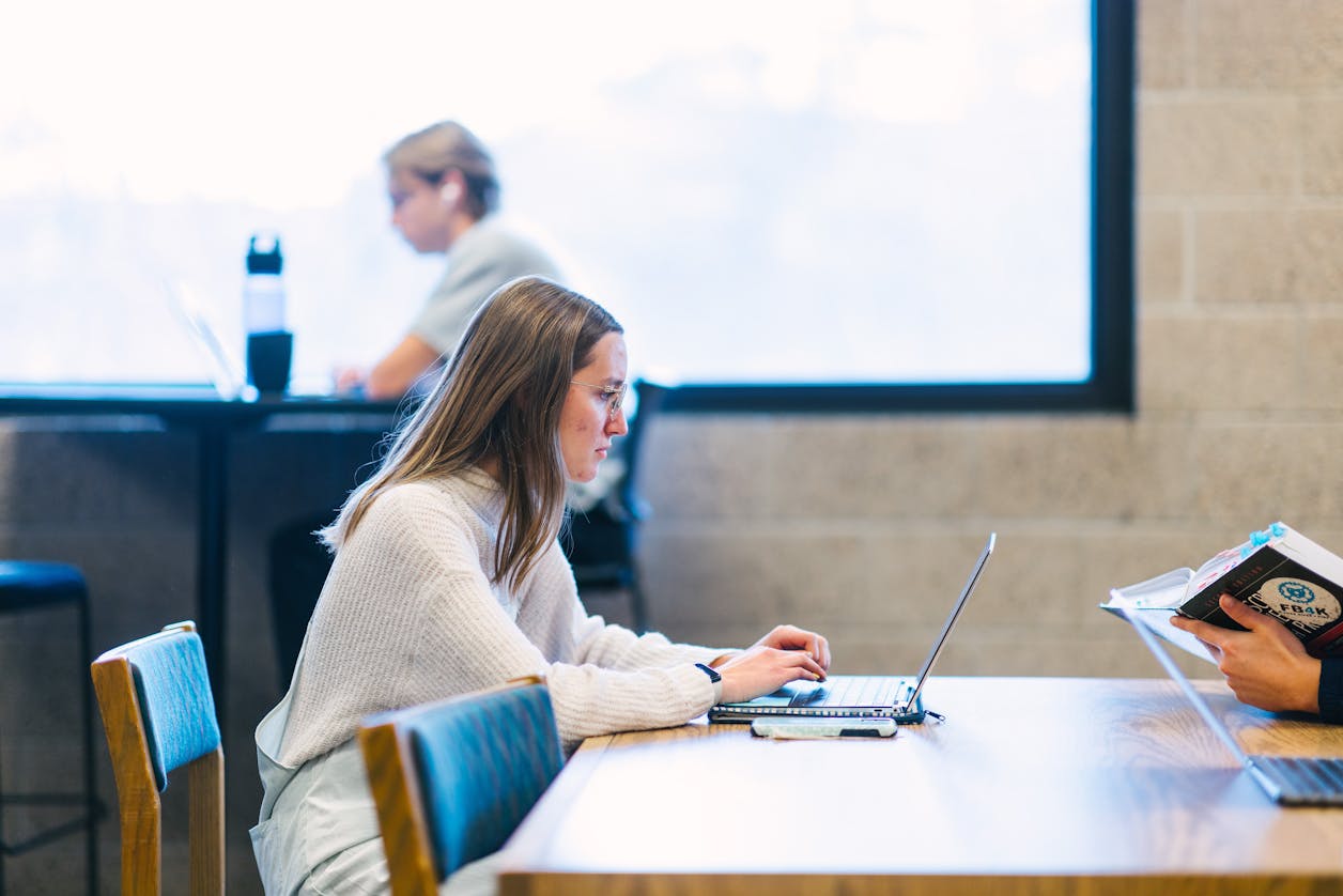 A student in a white sweater is intently working on her laptop in a bright study area. 