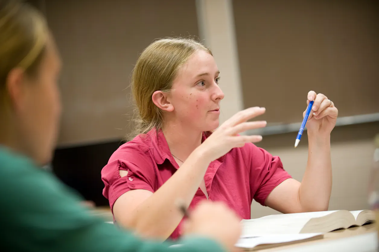 A student in a red shirt making a point during a classroom discussion, holding a pen.