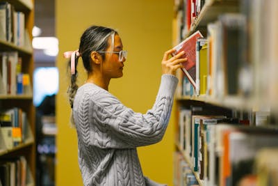 A student with a pink ribbon in her hair is selecting a book from the library shelves. 