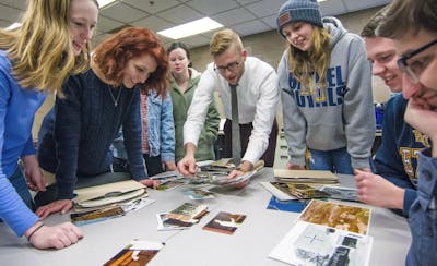 Students gather around a table full of photos while the professor presents them.