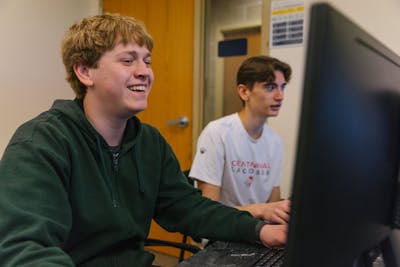 Two Bethel University students collaborating on a project, smiling and working on a desktop computer.