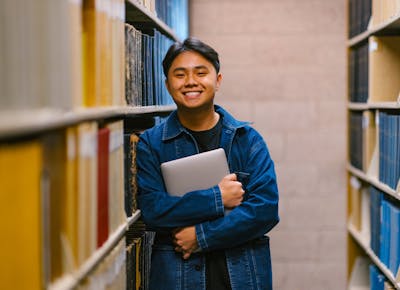 A smiling student casually leans against a library book shelf while holding 