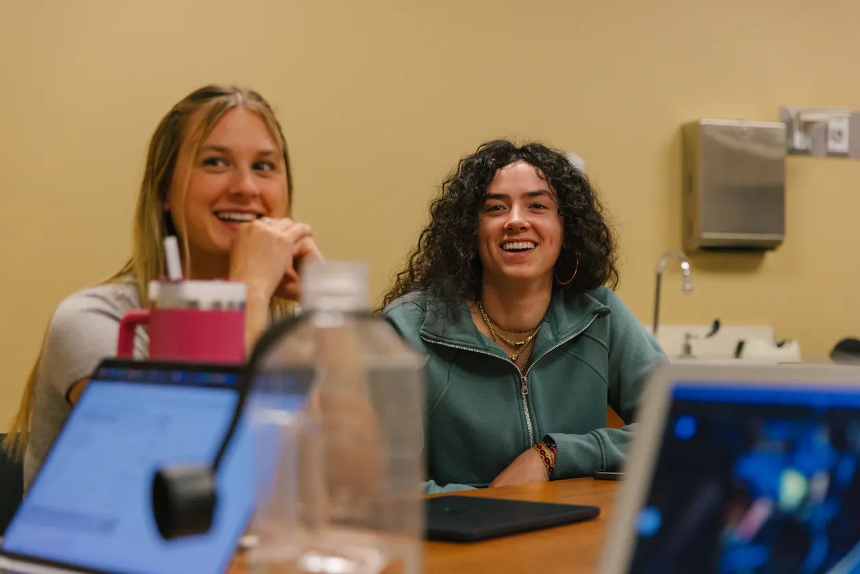 Two students smile as they listen to a classroom lecture.
