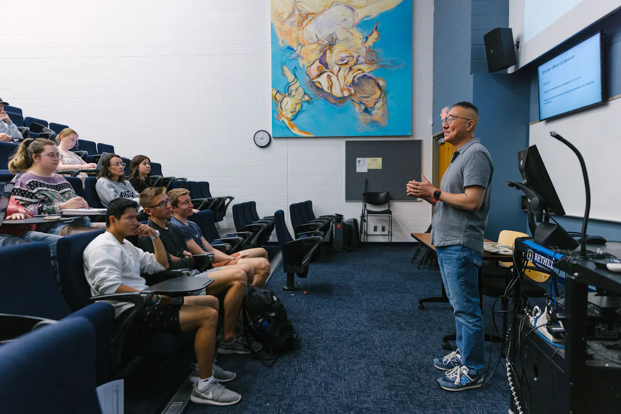 Professor lecturing to attentive students in a large classroom with vibrant artwork on the wall.