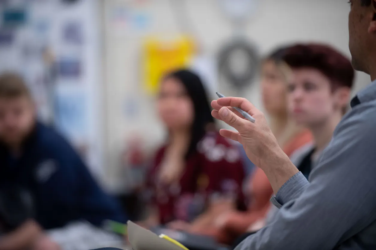 A professor's hand gestures during an engaging lecture, with students attentively listening in the background.