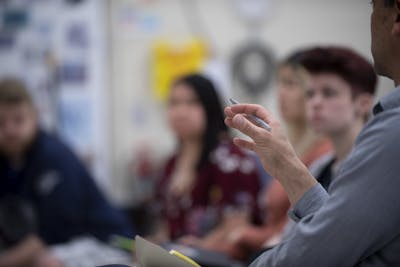 A professor's hand gestures during an engaging lecture, with students attentively listening in the background. 