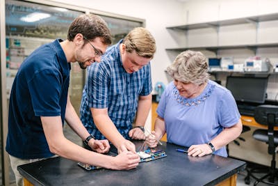 Two male students and a female professor collaborate on an electronics project in a lab.