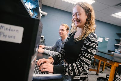 Two students smile while working on a laptop in a modern physics lab.