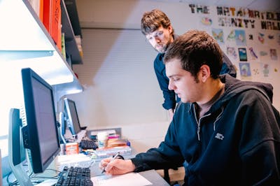 A professor assists a student with a computer assignment in a cozy office space.