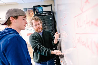 A professor in a green sweater explains concepts on a whiteboard to a student in a cap.