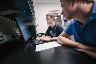 Two students work side by side on a computer, concentrating on their coding task.