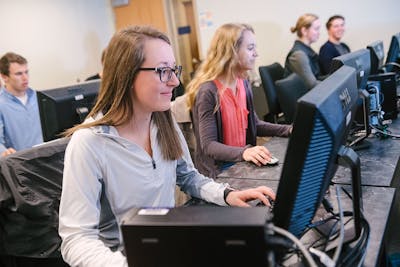 A group of students, including two females in front, work on their computers in a lab.