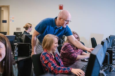 A professor in a blue shirt helps a student in a computer lab, both smiling.