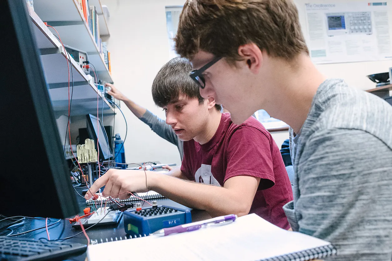 Two male students work on a technical project with circuits and notes in a lab.