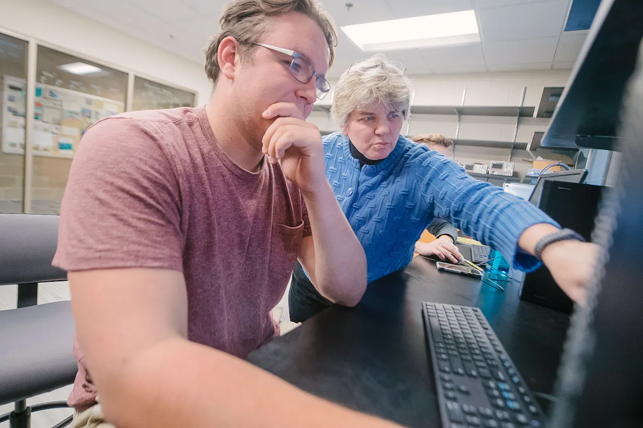 Professor in blue sweater guiding a student through computer work at Bethel University.