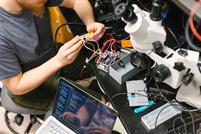 A student working on an electronic project, surrounded by wiring and a laptop, under a microscope.