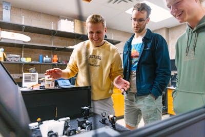 Three students excitedly discussing a physics experiment setup in a lab.