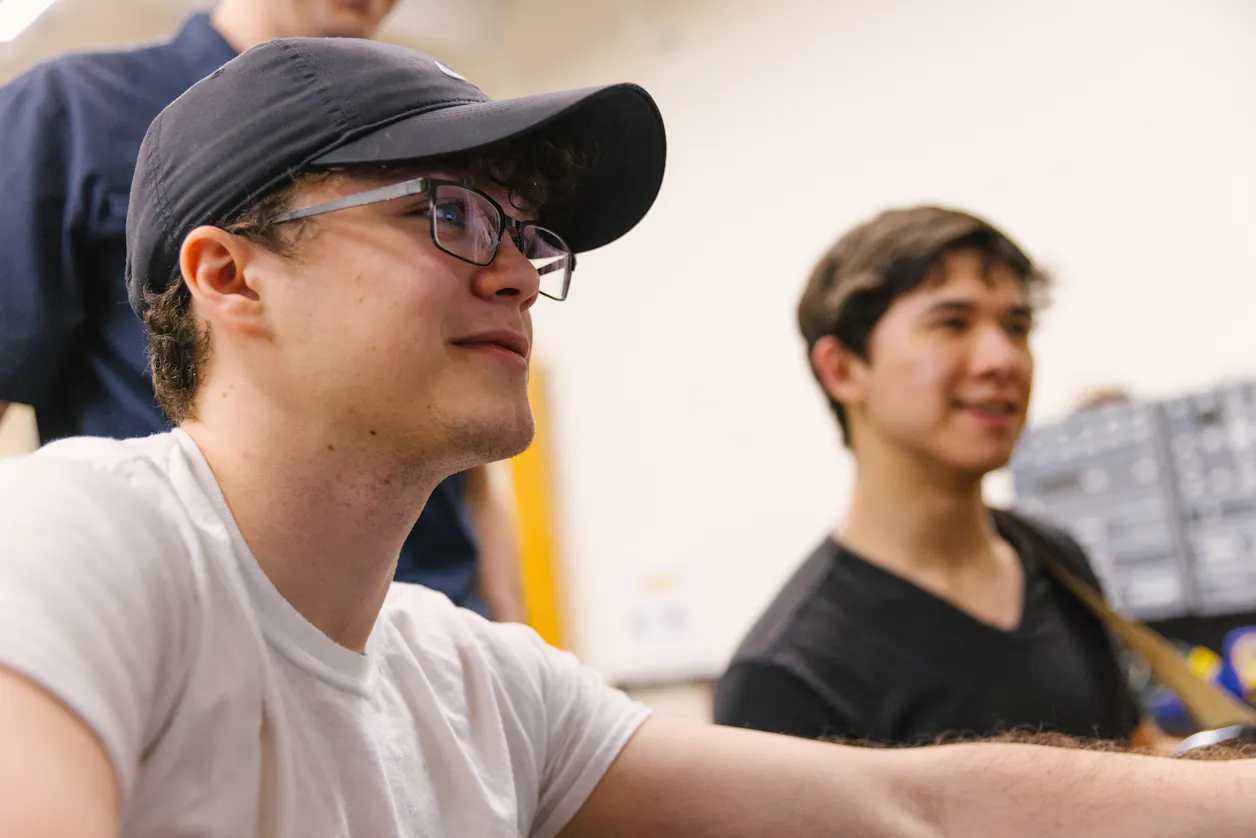 Students in a computer lab, one wearing glasses and a cap, focused on their work.