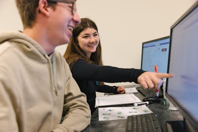 Student pointing at data on a computer screen as a smiling student looks on.