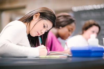 A Bethel University student concentrating on writing notes during a class, with classmates in the background. 