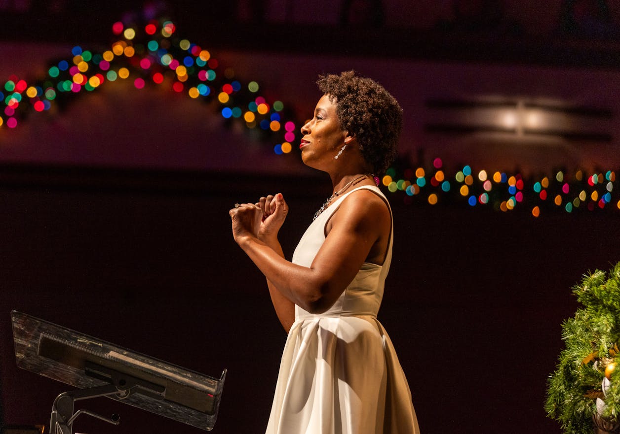 Woman in a white dress speaking passionately on stage with colorful lights in the background.