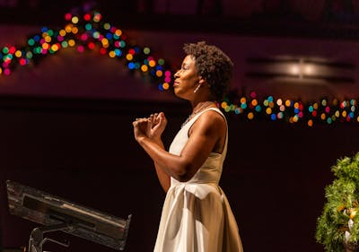 Woman in a white dress speaking passionately on stage with colorful lights in the background.