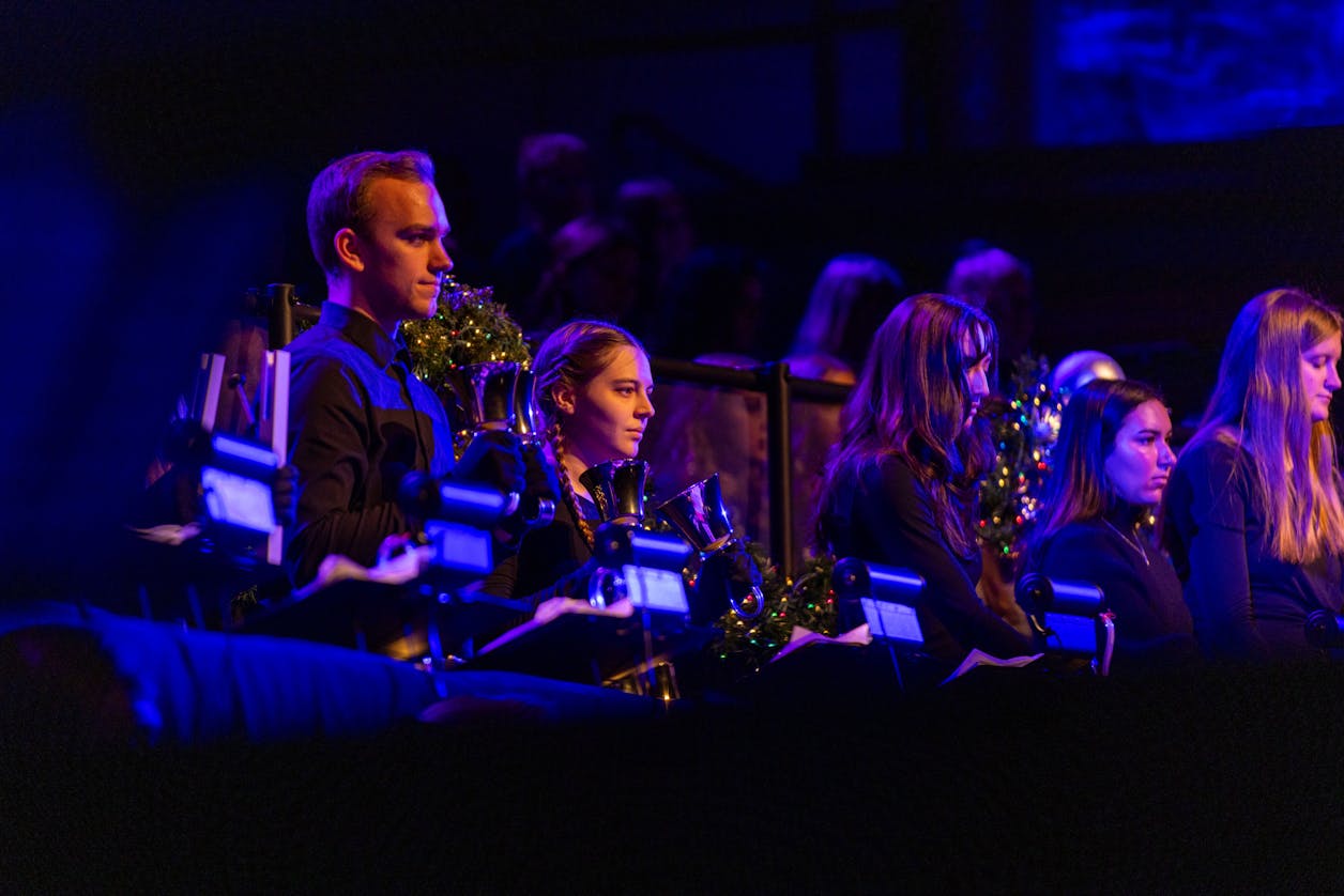 Musicians playing handbells during a performance, illuminated by blue stage lighting.