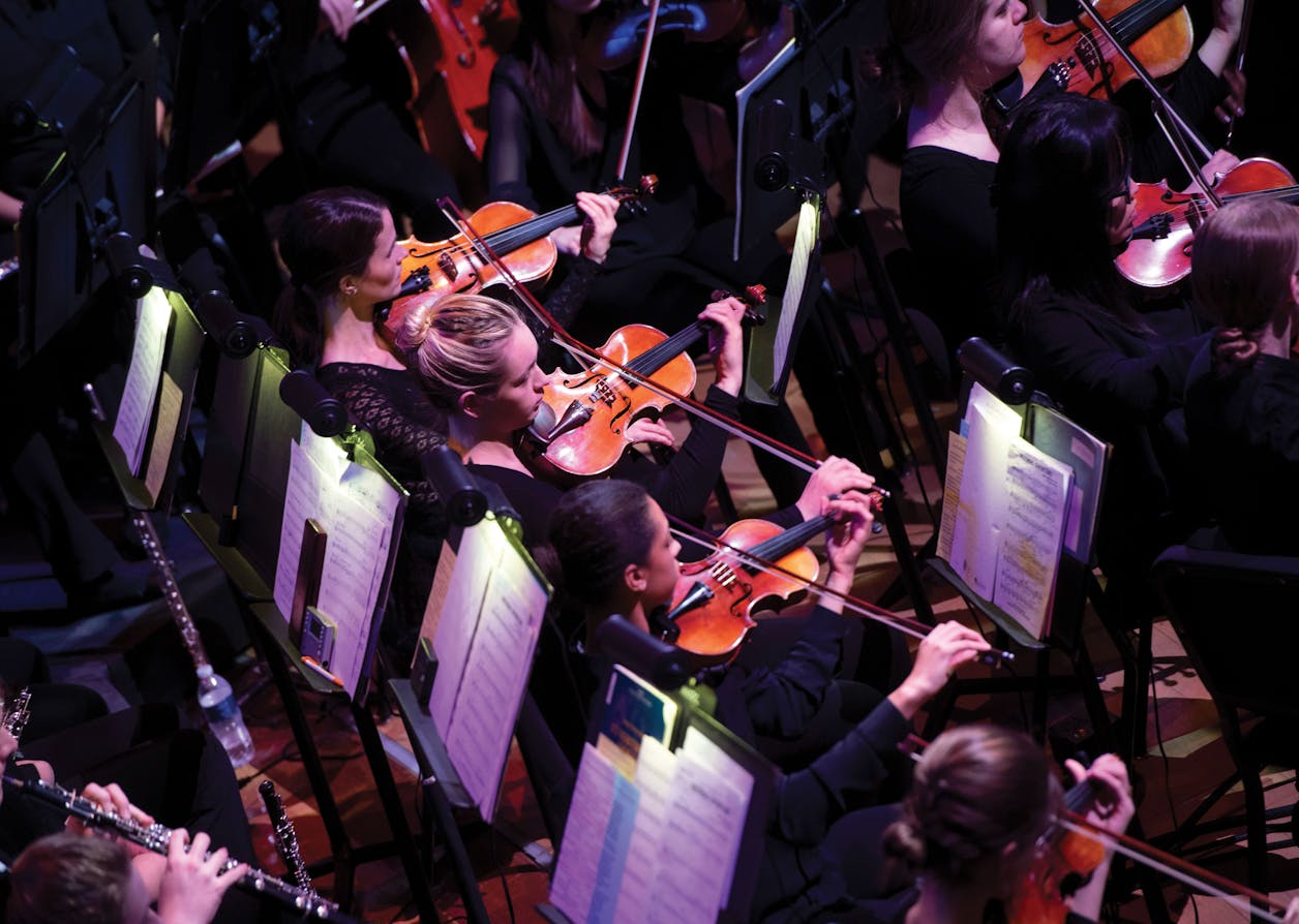 Violinists performing in an orchestra at Bethel University, captured from above with sheet music lit on stands.