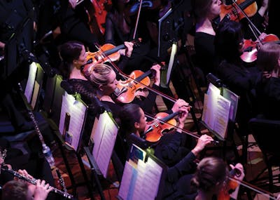 Violinists performing in an orchestra at Bethel University, captured from above with sheet music lit on stands.