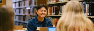 A man smiles at two other students while working at a computer with library books on shelves in the background. 