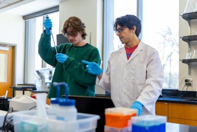 Two students collaborate in a Bethel University lab, one giving instructions while the other uses a pipette.