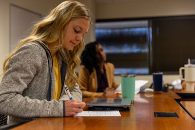 A focused student taking notes during a lecture, with another student in the background.