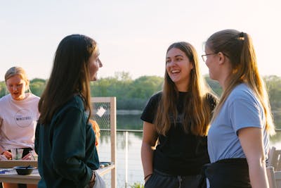 A group of smiling students talk outside near a lake.