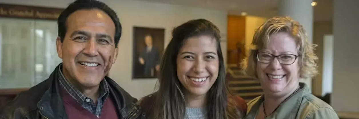 Smiling young woman standing between two adults, all looking at the camera in a warmly lit indoor setting.