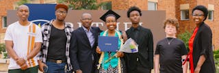 A smiling graduate in a cap and gown holds a diploma and flowers, surrounded by family in front of a Bethel University sign.