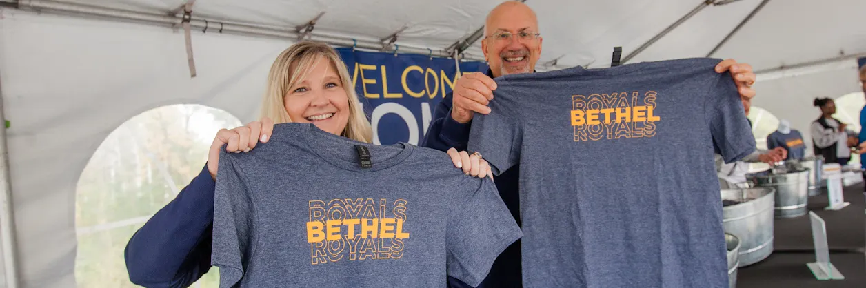Two smiling individuals hold up Bethel Royals T-shirts under a tent at a welcome event, with a "Welcome" sign in the background.
