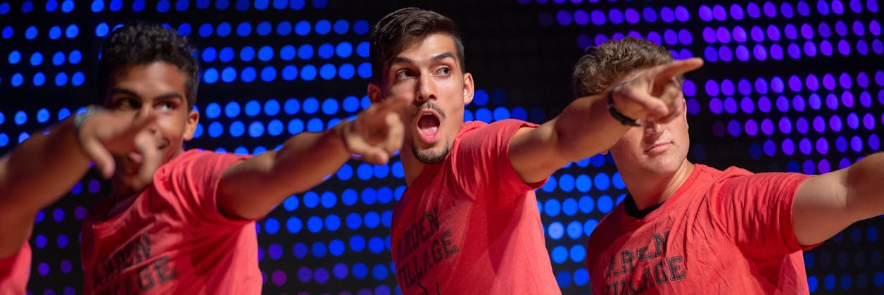 Three young men in red Arden Village shirts energetically point forward on a lit stage.
