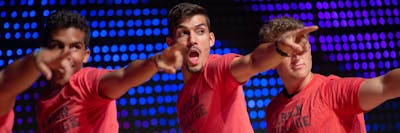 Three young men in red Arden Village shirts energetically point forward on a lit stage.