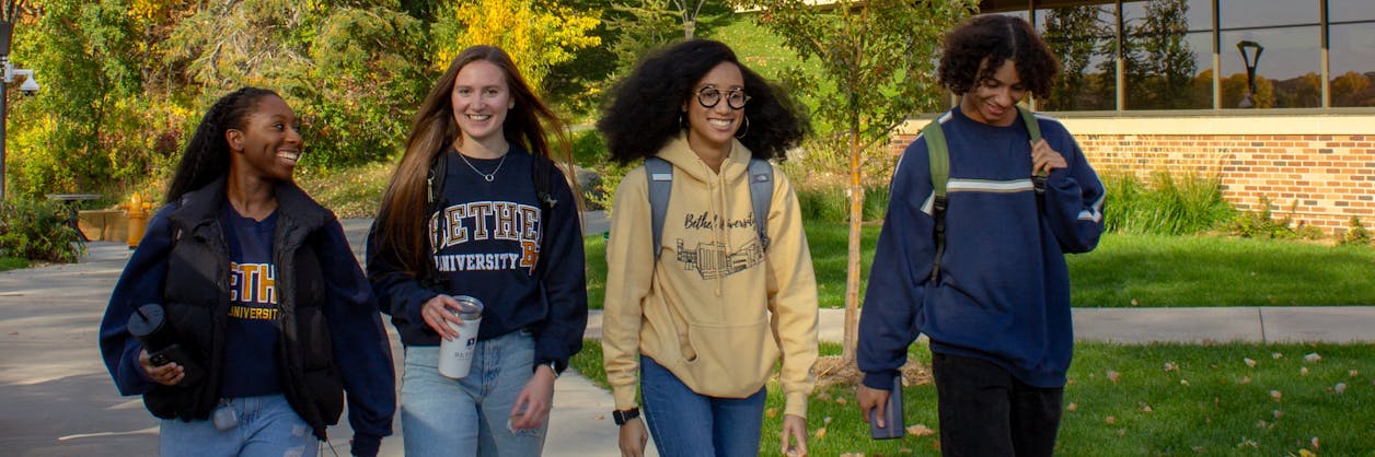 Four students smile as they walk together on Bethel's campus with changing leaves behind them. 