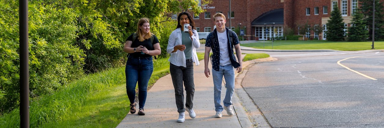  Three college students walking together on a tree-lined campus path, smiling and holding drinks.