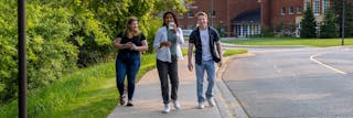  Three college students walking together on a tree-lined campus path, smiling and holding drinks.